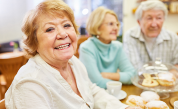 Oudere dame zit lachend aan tafel met tafelgenoten. 