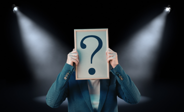 Spotlights gericht op persoon met bord met vraagteken voor z'n gezicht. 