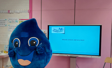Mascotte Druppie staat voor een TVscherm met een presentatie over de Nationale Kraanwaterdag